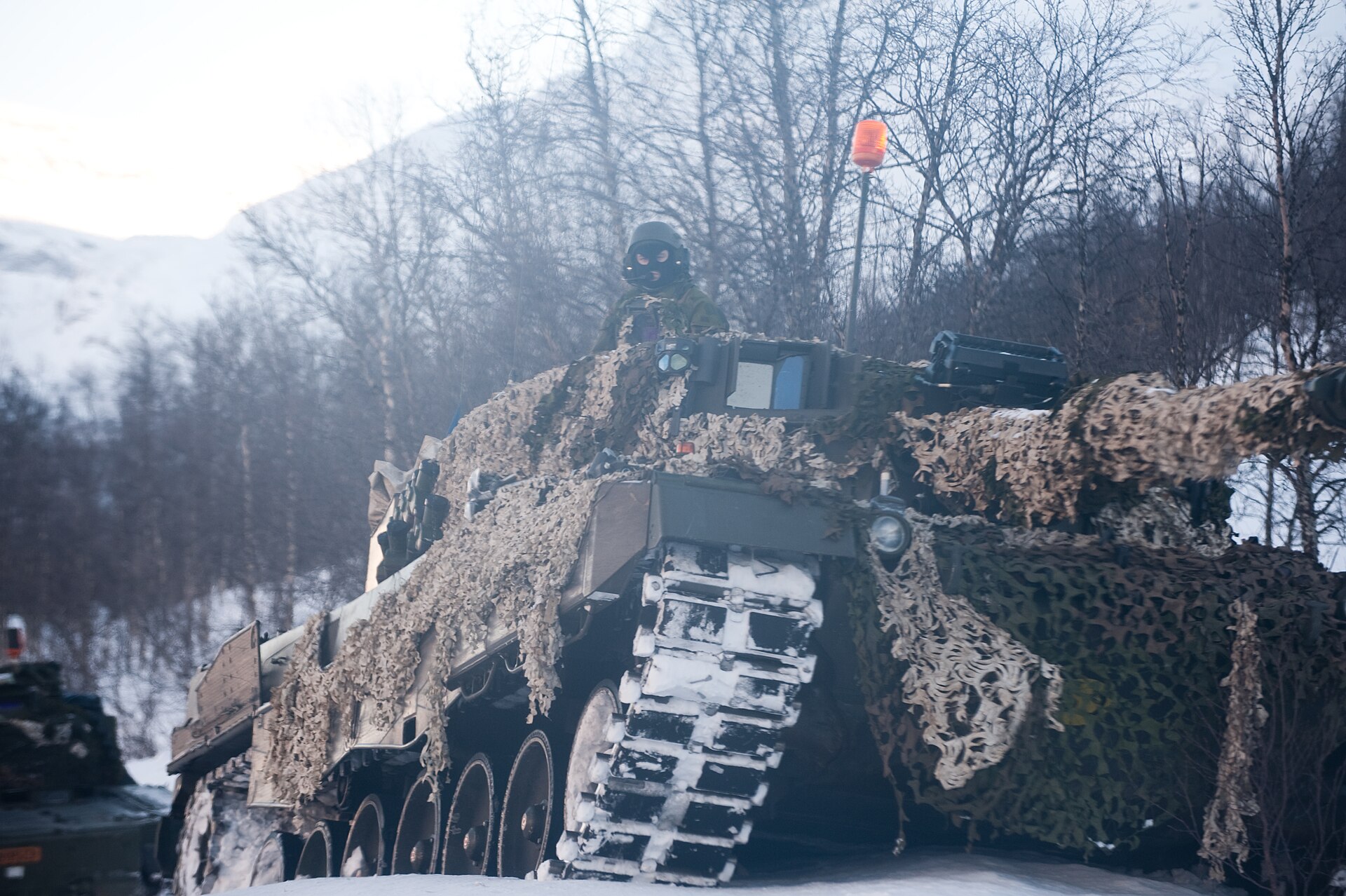 Un char norvégien recouvert d’un filet de camouflage dans un paysage enneigé, le chef de char en observation