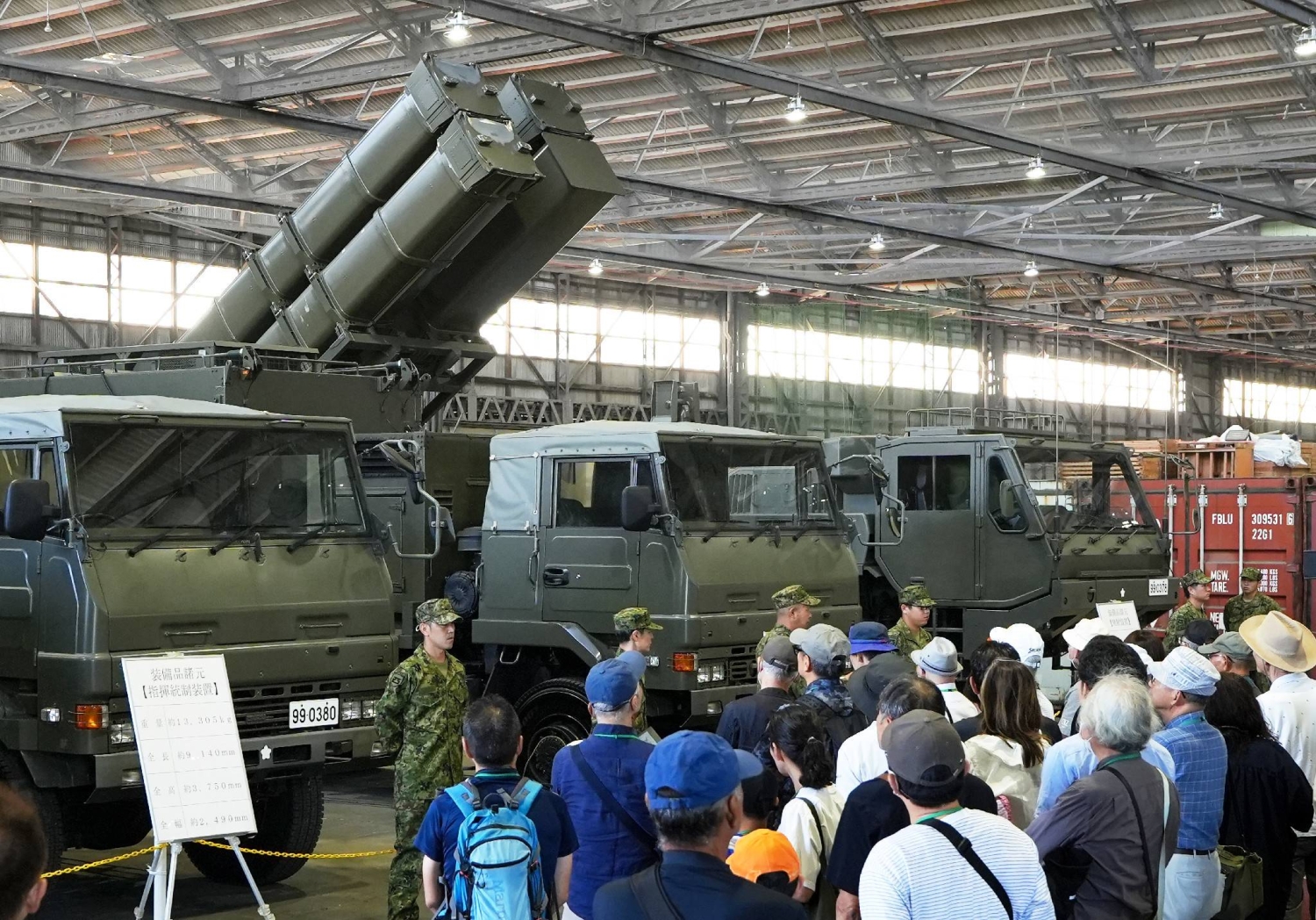 Visiteurs civils rassemblés devant les véhicules du système Type 25 dans un hangar du camp de Kengun, lanceur aux caissons relevés à l'arrière-plan